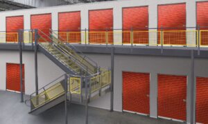 A clean, well-lit self-storage facility with red roll-up doors and a central staircase with yellow handrails, leading to upper-level units.