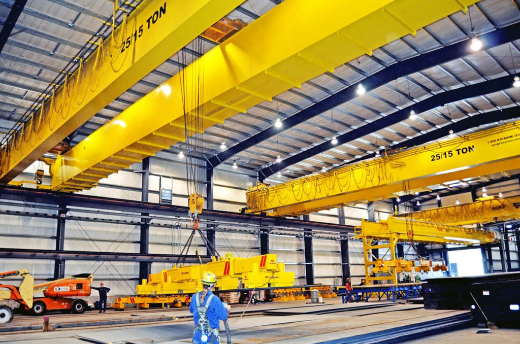 Warehouse Bright warehouse scene: two yellow overhead cranes (one "25/5 TON") lifting equipment, workers present. Transform Your Workflow with Advanced Hoist and Crane Systems.